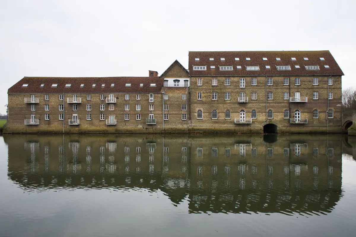 A long brick building beside water, its windows reflected in the surface.